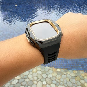 Pode incluir: Smartwatch preto com moldura dourada e pulseira de silicone preta. O relógio é mostrado em um pulso, com a tela voltada para o espectador. O relógio tem um mostrador retangular e um design moderno.