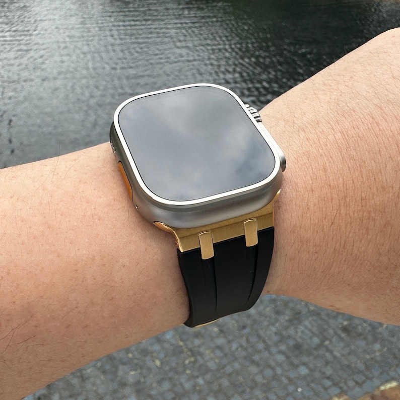 Może przedstawiać: Srebrny i złoty zegarek Apple Watch z czarnym paskiem. Tarcza zegarka jest czarna, a pasek wykonany jest z wytrzymałego materiału.