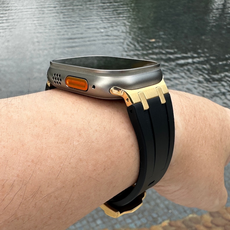 Może przedstawiać: Srebrny smartwatch z czarnym paskiem z gumy i złotymi akcentami. Zegarek ma okrągłą tarczę i mały pomarańczowy przycisk z boku.
