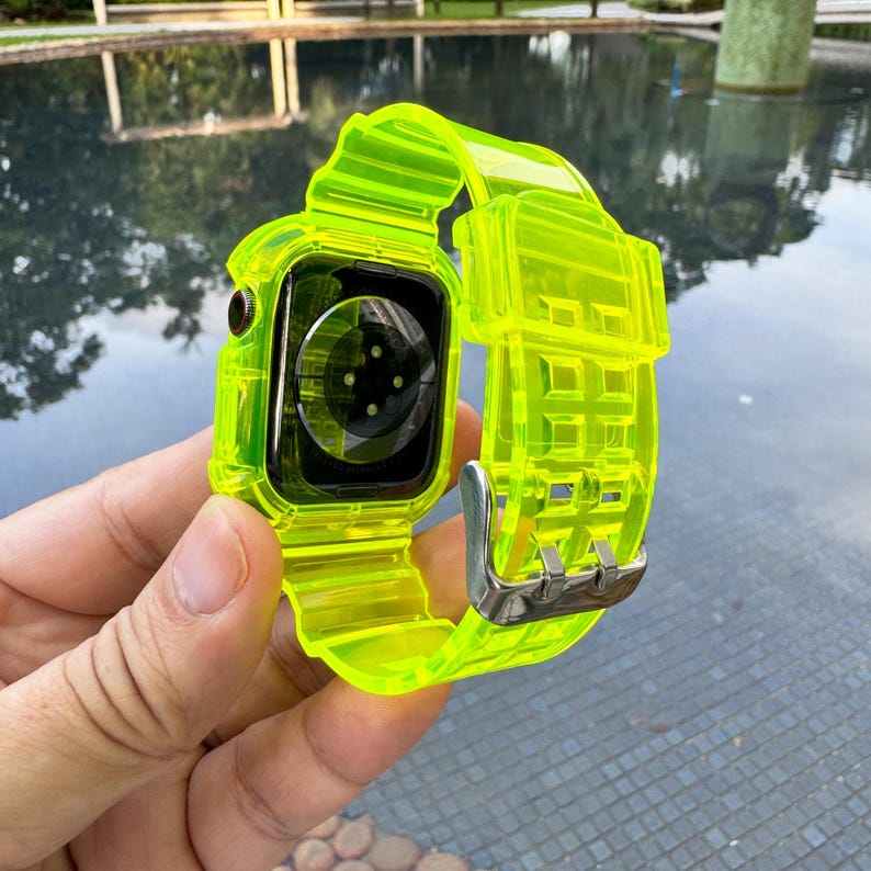 May include: A neon green smartwatch with a transparent band and case. The watch has a silver buckle and a black screen. The band is made of a flexible, ribbed material. The watch is held in a hand.