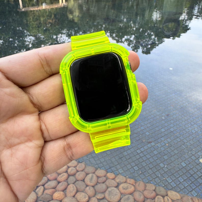 May include: A neon green smartwatch case and band. The watch face is black. The band is a transparent, flexible material. The watch is held in a hand against a background of water and stones.
