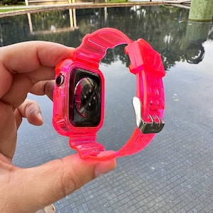 May include: A bright pink clear silicone watch band for an Apple Watch. The band is made of a flexible, transparent material and has a secure clasp.