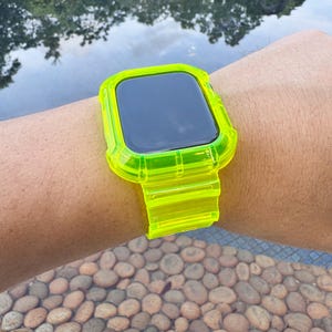 May include: A neon green watch case and band are shown on a wrist. The watch face is black. The band and case are made of a transparent, bright green material. The watch is shown against a background of water and pebbles.