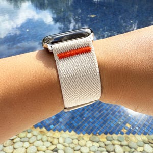Può includere: Un cinturino beige per Apple Watch con una striscia arancione brillante. Il cinturino è realizzato in materiale tessuto ed è attaccato a una cassa dell'orologio argentata. L'orologio è su un polso, con uno sfondo blu e di pietra.