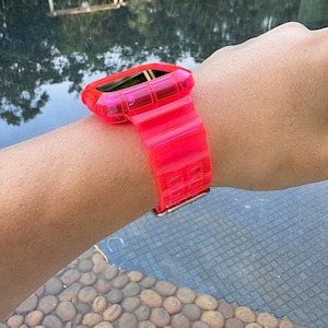 May include: A bright pink clear plastic watch band with a matching case. The band has a grid pattern and is worn on a person's wrist.