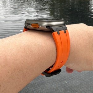Puede incluir: Un reloj inteligente plateado y negro con una banda de silicona naranja. El reloj est&aacute; en la mu&ntilde;eca de una persona.