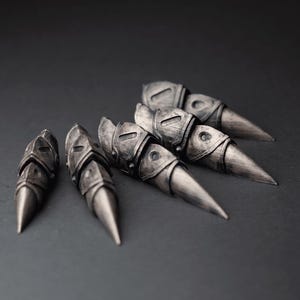 May include: A set of five silver-colored finger armor pieces. Each piece has a pointed tip and a detailed, armored design. The pieces are arranged on a dark gray surface.