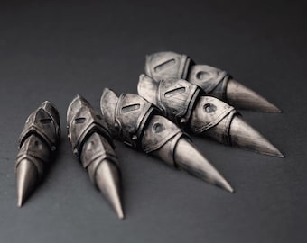 May include: A set of five silver-colored finger armor pieces. Each piece has a pointed tip and a detailed, armored design. The pieces are arranged on a dark gray surface.