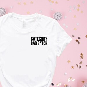 May include: White t-shirt with the text "CATEGORY BAD B*TCH" printed on the front. The shirt is surrounded by silver and gold stars on a pink background.