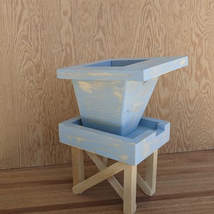 May include: A light blue wooden planter with a square top and a flared base. The planter is on a wooden stand with an X-shaped base.