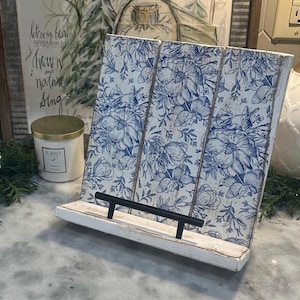 Könnte beinhalten: Ein Kochbuchständer aus Holz mit weißem Finish und einem blauen Blumenmuster auf drei vertikalen Paneelen. Der Ständer verfügt über eine horizontale schwarze Metallstange an der Vorderkante, um Bücher oder Tablets aufrecht zu halten.
