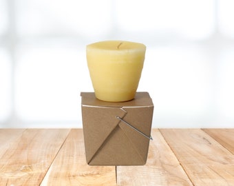 May include: A yellow candle sits on top of a brown paper takeout container. The container has a wire handle.