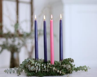 May include: Four candles, three purple and one pink, are arranged in a wreath of green foliage. The candles are lit and the flames are visible.
