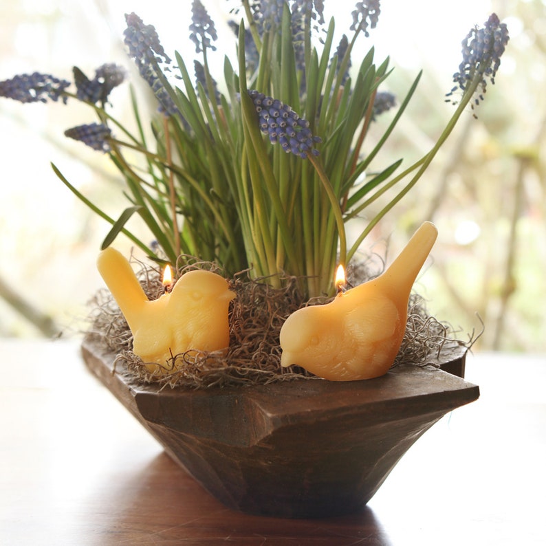 May include: Two small bird-shaped candles are nestled in a wooden bowl with a bed of moss. The candles are lit and the bowl is filled with a bouquet of purple and blue grape hyacinths.
