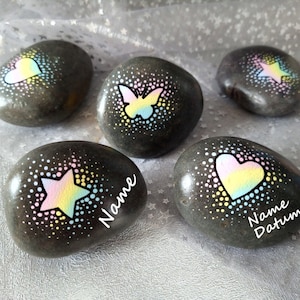 Könnte beinhalten: Fünf bemalte graue Steine mit regenbogenfarbenen Designs. Die Designs beinhalten ein Herz, einen Schmetterling, einen Stern und zwei Herzen. Die Steine haben die Aufschriften "Name" und "Name Datum" auf sich.