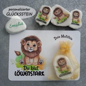 Könnte beinhalten: Eine Sammlung personalisierter Glückssteine mit einer Löwenjungen-Illustration. Das Set enthält Steine mit Texten wie "Du bist Löwenstark" und "Emilia", zusammen mit einer Karte und einem kleinen Beutel.