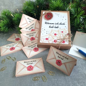 Könnte beinhalten: Dekorative Papierumschläge mit Wachssiegel und dem deutschen Text "Warum ich dich lieb hab...". Sie sind um eine kleine braune Schachtel mit passendem Design angeordnet und mit Bindfaden verschnürt. Goldene Büroklammern sind verstreut.