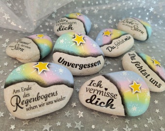 May include: A collection of seven smooth, oval-shaped stones painted with pastel rainbow gradients, white stars, and yellow star accents. Each stone features black German text including phrases like Unvergessen, Du fehlst mir, and Wir vermissen dich, arranged on a surface.