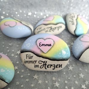 Könnte beinhalten: Bemalte herzförmige Steine mit dem Text "For immer im Herzen" und dem Namen "Emma" auf einem der Steine. Die Steine sind mit Pastellfarben und Glitzer verziert.
