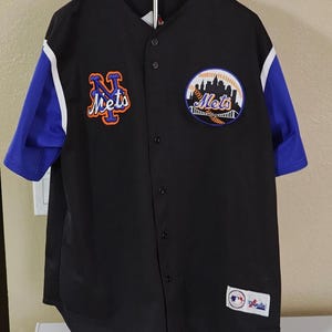 May include: Black baseball jersey with blue sleeves and white accents. Features the Mets logo on the chest and left sleeve. The jersey has a button-up front and a Majestic brand tag.