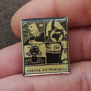 May include: A rectangular metal pin featuring a black and gold graphic design divided into four panels. The illustrations depict a person with a stethoscope, a globe, a smiling person, and a face with a head covering. The text reads MEDICOS SIN FRONTERAS.