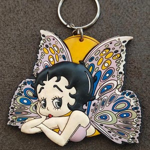 May include: A Betty Boop keychain featuring the cartoon character with butterfly wings. The keychain has a silver ring and a yellow sun-like background. The wings are detailed with blue, purple, and yellow patterns, and the character is in a pink bra.