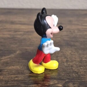 May include: A small Mickey Mouse figurine. The figure has a black head with large ears, a pink face, and a black nose. It wears a blue shirt, red pants, and yellow shoes. Its white gloved hands are raised.