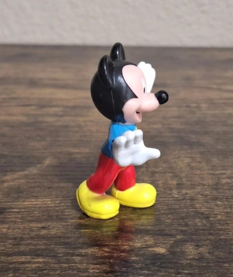 May include: A small Mickey Mouse figurine. The figure has a black head with large ears, a pink face, and a black nose. It wears a blue shirt, red pants, and yellow shoes. Its white gloved hands are raised.