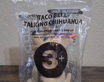 May include: A plastic bag containing a Taco Bell talking Chihuahua plush toy. The toy is brown and white with a purple collar and a 3+ age recommendation. The bag has text that reads "Taco Bell Talking Chihuahua" and "For children ages 3 and up".