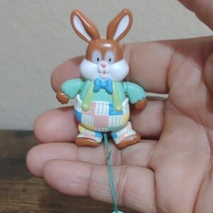 May include: A small, brown and white bunny figurine with green and blue checkered pants and a green bow tie. The bunny is attached to a green string with a white circle at the end.