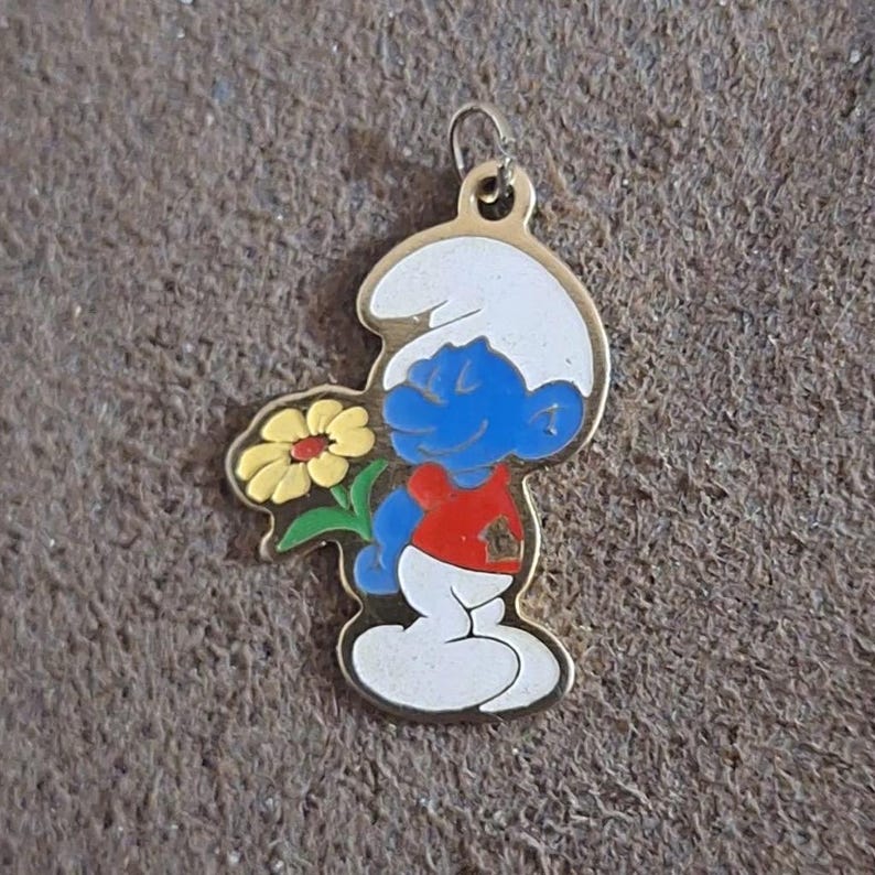 May include: A gold-toned Smurf charm holding a yellow flower with green leaves. The Smurf is blue with a white hat, red shirt, and white pants. The charm has a loop at the top for attachment.