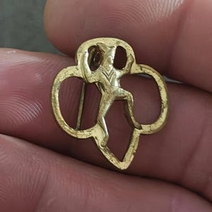 May include: A gold-colored pin featuring the Girl Scout trefoil emblem. The pin has a stylized figure in the center, with a pin back for attachment. The design is a classic symbol of the Girl Scouts.