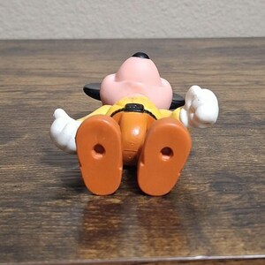 Puede incluir: Una figura de juguete de Goofy, acostado boca arriba. La figura tiene una cara rosa, orejas negras y una camisa amarilla con pantalones naranjas. Se ven guantes blancos y pies naranjas. La figura est&aacute; sobre una superficie de madera.