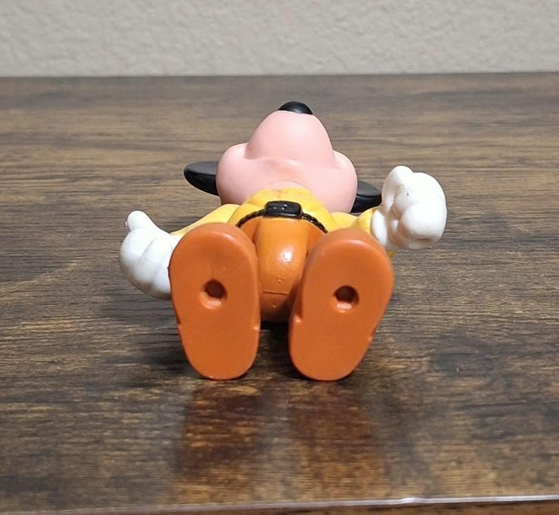 Puede incluir: Una figura de juguete de Goofy, acostado boca arriba. La figura tiene una cara rosa, orejas negras y una camisa amarilla con pantalones naranjas. Se ven guantes blancos y pies naranjas. La figura est&aacute; sobre una superficie de madera.