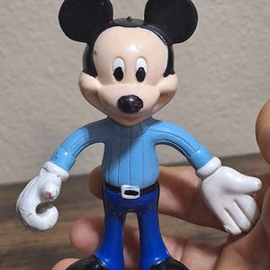 Puede incluir: Una figura de Mickey Mouse con orejas negras, nariz negra y tez clara. Viste un su&eacute;ter azul claro, pantalones azul oscuro, zapatos negros y guantes blancos. La figura est&aacute; sujeta por una mano.