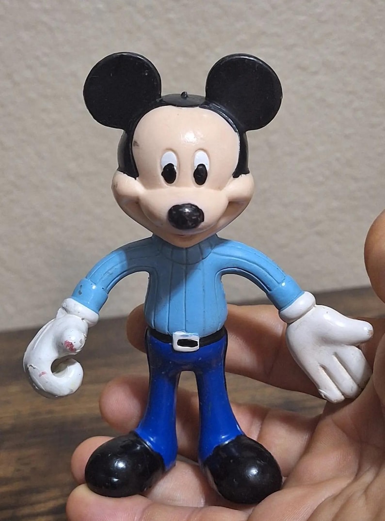 Puede incluir: Una figura de Mickey Mouse con orejas negras, nariz negra y tez clara. Viste un su&eacute;ter azul claro, pantalones azul oscuro, zapatos negros y guantes blancos. La figura est&aacute; sujeta por una mano.