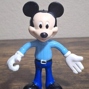 Puede incluir: Una figura de Mickey Mouse. La figura tiene una cabeza negra con orejas grandes, una cara blanca y una nariz negra. Viste un su&eacute;ter azul claro, pantalones azules, zapatos negros y guantes blancos. La figura est&aacute; de pie con los brazos extendidos.