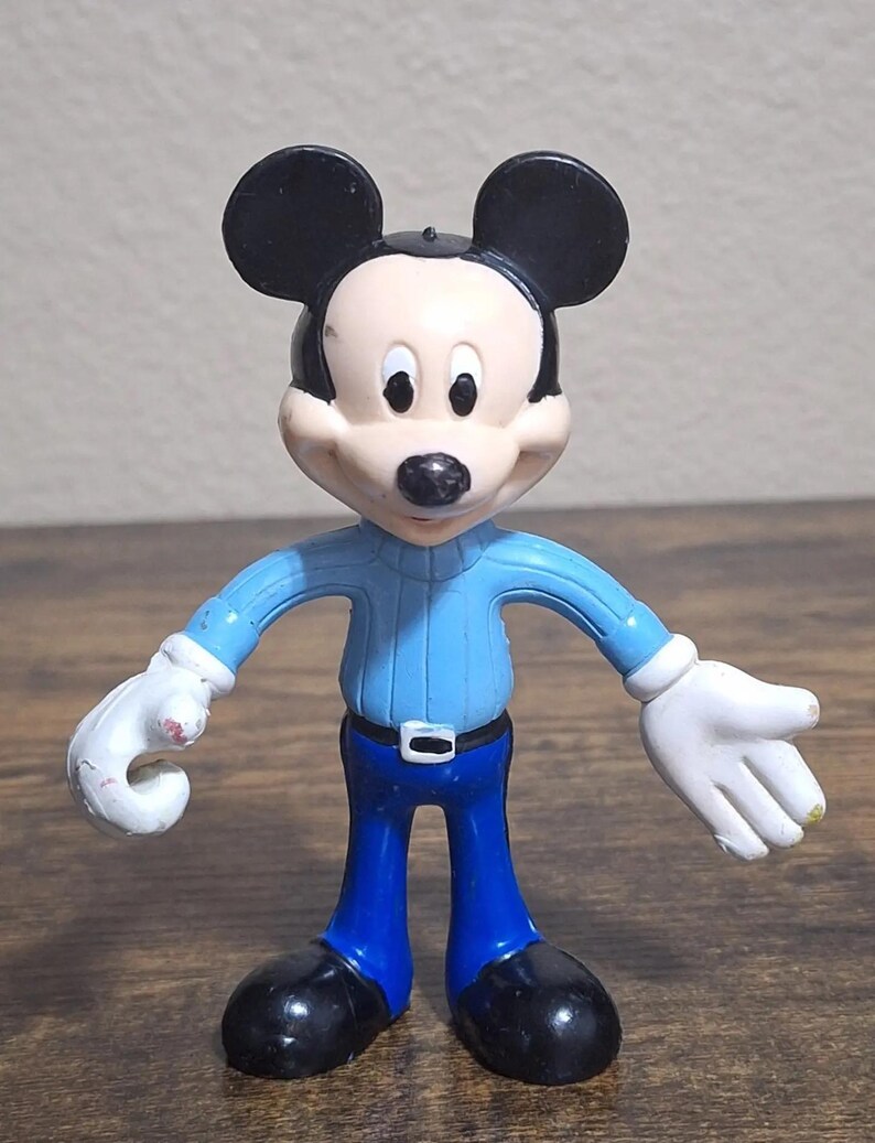 Puede incluir: Una figura de Mickey Mouse. La figura tiene una cabeza negra con orejas grandes, una cara blanca y una nariz negra. Viste un su&eacute;ter azul claro, pantalones azules, zapatos negros y guantes blancos. La figura est&aacute; de pie con los brazos extendidos.