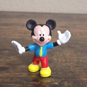 May include: A small Mickey Mouse figurine. Mickey is wearing a blue shirt, red pants, yellow shoes, and a green tie. His hands are white, and his ears are black. The figurine is standing on a brown surface.