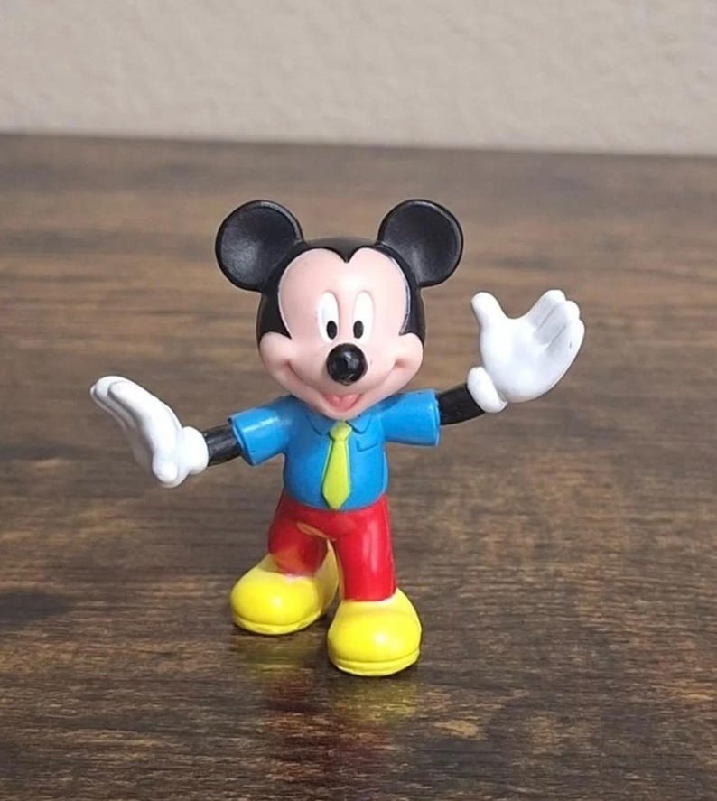 May include: A small Mickey Mouse figurine. Mickey is wearing a blue shirt, red pants, yellow shoes, and a green tie. His hands are white, and his ears are black. The figurine is standing on a brown surface.
