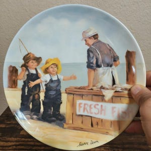 May include: Decorative plate featuring a seaside scene with two children in overalls and hats, interacting with a fishmonger. A wooden stand displays "FRESH FISH". The plate is signed "Jeanne Down".