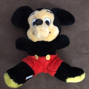 May include: A black and red plush Mickey Mouse toy with yellow ears and a big smile. The toy is sitting on a brown surface.