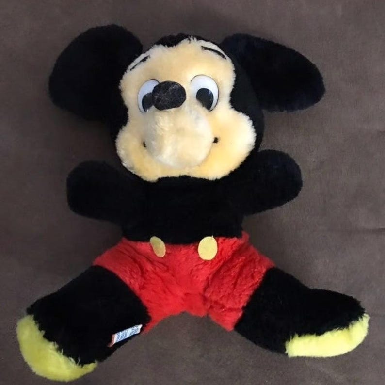 May include: A black and red plush Mickey Mouse toy with yellow ears and a big smile. The toy is sitting on a brown surface.