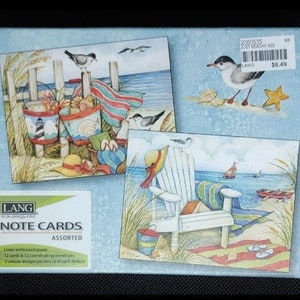 May include: A box of assorted note cards featuring two beach-themed designs. The first design shows a beach scene with a lighthouse, buckets, and seagulls. The second design shows a white Adirondack chair on a beach with a sailboat in the distance. The box is labeled "Lang Note Cards Assorted" and includes 12 cards and 12 coordinating envelopes.