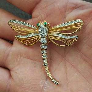 May include: A gold-toned dragonfly brooch with clear and green gemstones. The dragonfly's wings are outlined with clear stones, and the body is covered in clear stones. The eyes are small green stones.