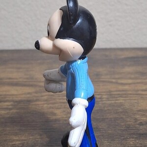 Puede incluir: Una figura de Mickey Mouse de perfil. La figura tiene cabeza y orejas negras, cara y nariz de color claro, camisa azul, guantes blancos y pantalones azules. La figura est&aacute; sobre una superficie de madera.