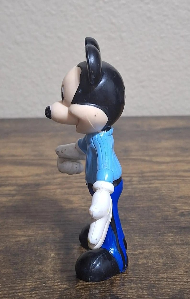 Puede incluir: Una figura de Mickey Mouse de perfil. La figura tiene cabeza y orejas negras, cara y nariz de color claro, camisa azul, guantes blancos y pantalones azules. La figura est&aacute; sobre una superficie de madera.