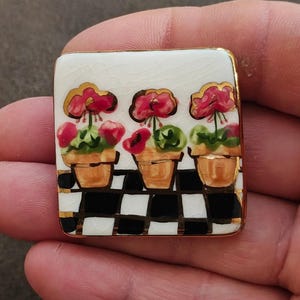 May include: Square ceramic brooch with a gold trim. The brooch features a design of three potted flowers with red blooms and green leaves, set against a black and white checkered pattern. The background is white.