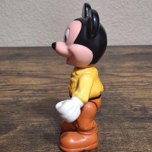 Puede incluir: Una figura vintage de Mickey Mouse. La figura tiene una cabeza negra con orejas grandes, una cara rosada y una camisa amarilla. Lleva pantalones naranjas, zapatos grandes naranjas y guantes blancos. El juguete est&aacute; sobre una superficie de madera.