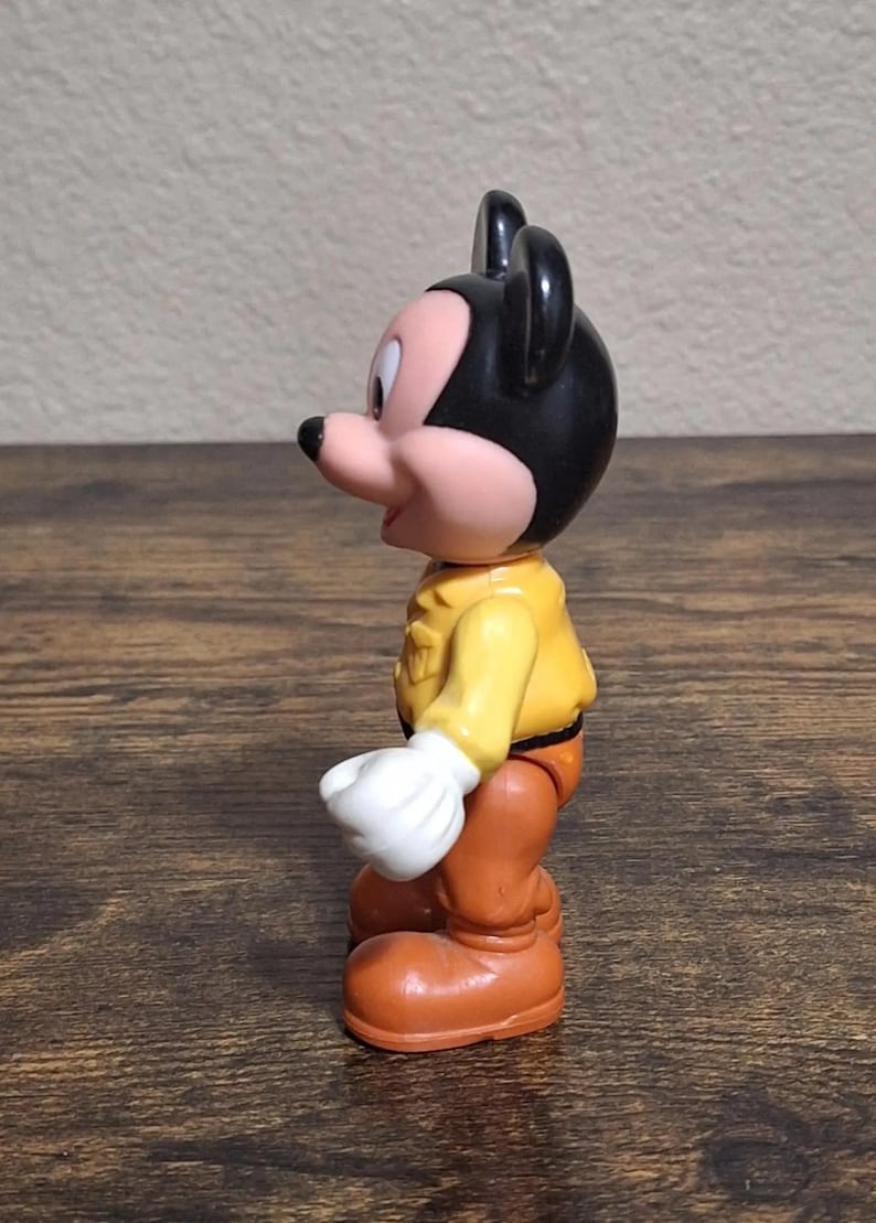 Puede incluir: Una figura vintage de Mickey Mouse. La figura tiene una cabeza negra con orejas grandes, una cara rosada y una camisa amarilla. Lleva pantalones naranjas, zapatos grandes naranjas y guantes blancos. El juguete est&aacute; sobre una superficie de madera.